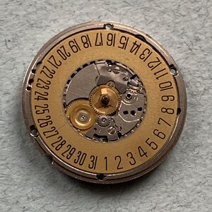 May include: A close-up of a watch movement with a gold-colored dial and silver-colored gears. The dial features the numbers 1 through 31, indicating the days of the month. The intricate mechanical components are visible.