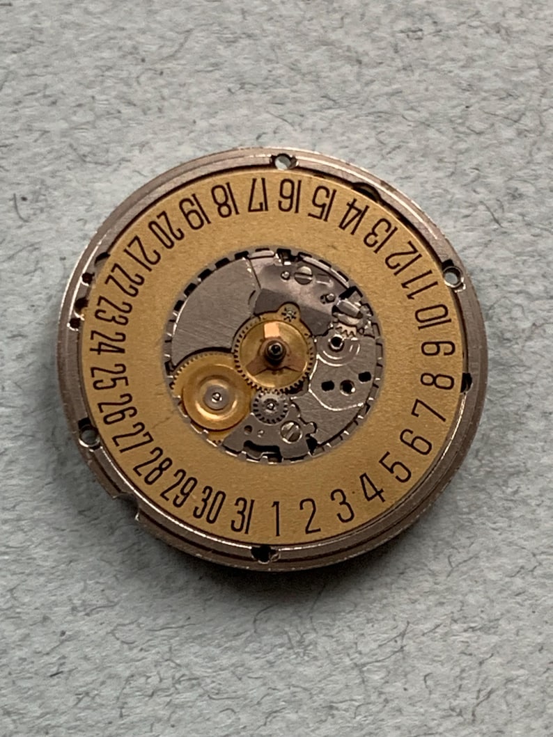 May include: A close-up of a watch movement with a gold-colored dial and silver-colored gears. The dial features the numbers 1 through 31, indicating the days of the month. The intricate mechanical components are visible.