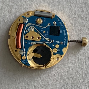 May include: A close-up of a watch movement with a gold-colored frame and a blue circuit board. The board has several components, including a battery, a coil, and the text "UNADJUSTED SEVEN 7 JEWELS". A screw with a gold-colored head is visible.
