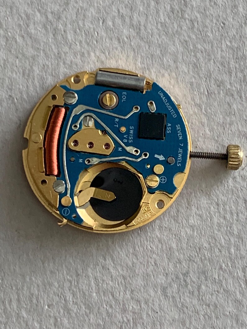 May include: A close-up of a watch movement with a gold-colored frame and a blue circuit board. The board has several components, including a battery, a coil, and the text "UNADJUSTED SEVEN 7 JEWELS". A screw with a gold-colored head is visible.