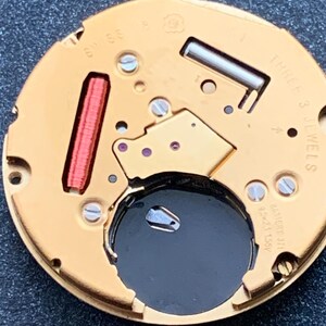 May include: Close-up of a watch movement with gold-colored components. The watch mechanism features a red coil, screws, and the text "SWISS" and "THREE 3 JEWELS". A black battery is visible in the center.