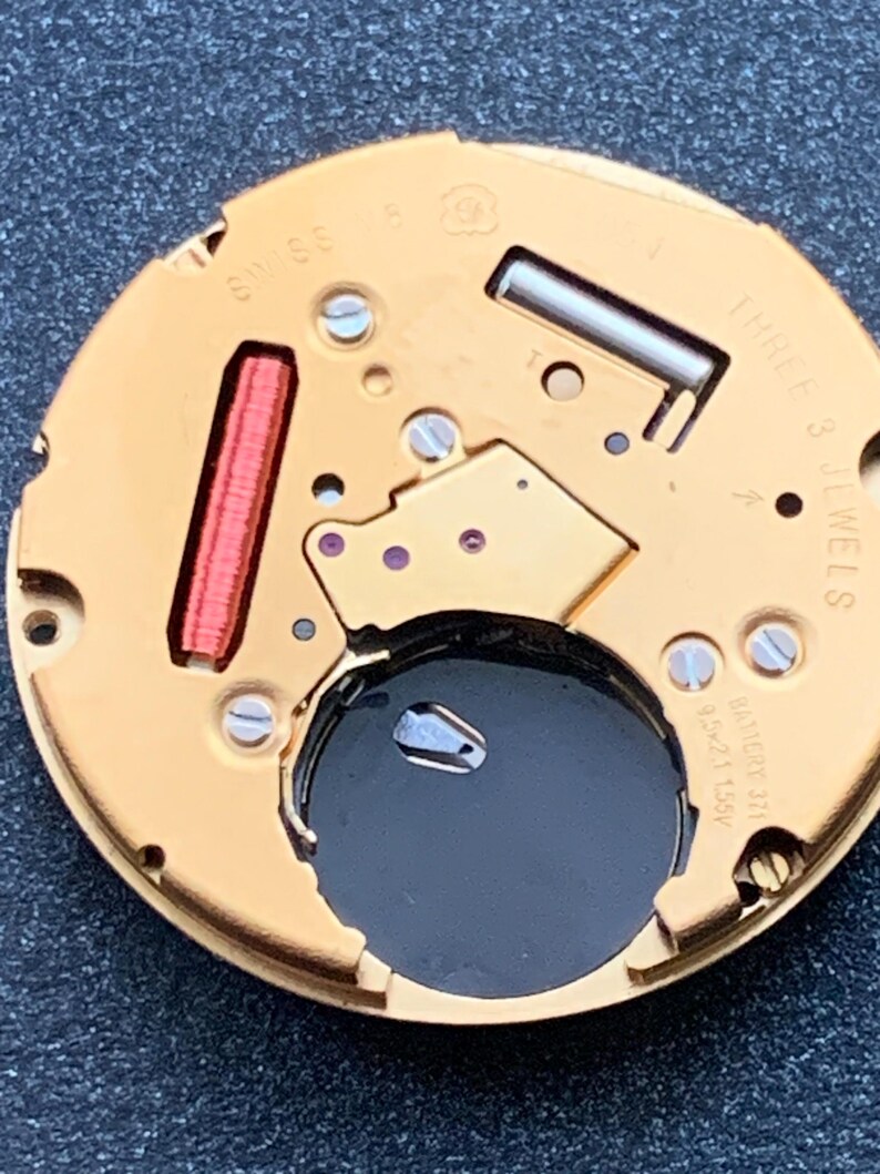 May include: Close-up of a watch movement with gold-colored components. The watch mechanism features a red coil, screws, and the text "SWISS" and "THREE 3 JEWELS". A black battery is visible in the center.