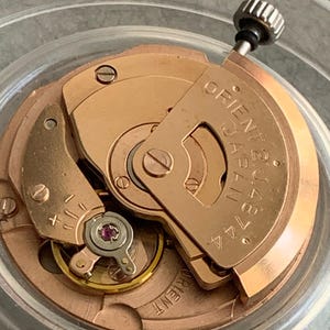 Puede incluir: Primer plano de un movimiento de reloj con engranajes y componentes dorados. El texto "ORIENT JAPAN 21J 46744" es visible en una placa met&aacute;lica. Una peque&ntilde;a corona negra en la parte superior.