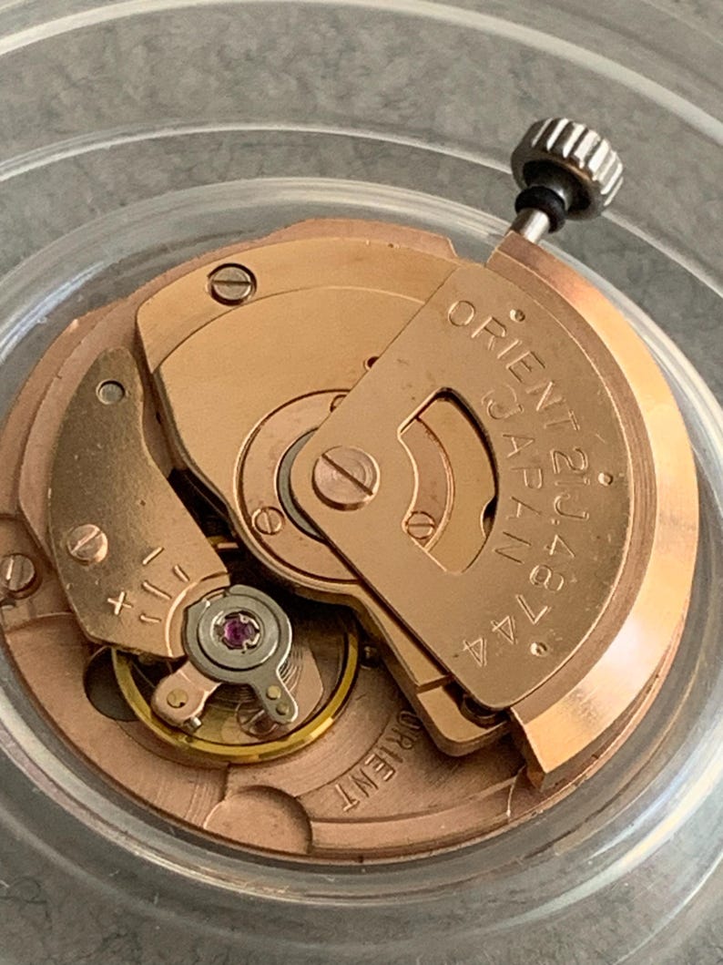 Puede incluir: Primer plano de un movimiento de reloj con engranajes y componentes dorados. El texto "ORIENT JAPAN 21J 46744" es visible en una placa met&aacute;lica. Una peque&ntilde;a corona negra en la parte superior.