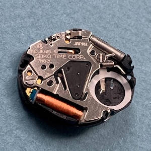 K&ouml;nnte beinhalten: Nahaufnahme eines Seiko-Uhrwerks. Der silberfarbene Mechanismus tr&auml;gt die Aufschrift "NO JEWELS", "SEIKO TIME CORP." und "7N82C". Eine kupferfarbene Spule und schwarze Komponenten sind sichtbar. Das Uhrwerk befindet sich vor blauem Hintergrund.
