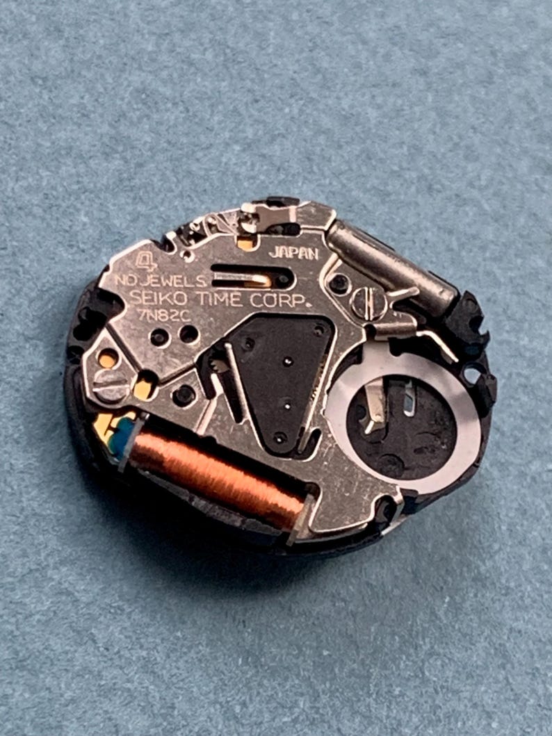 K&ouml;nnte beinhalten: Nahaufnahme eines Seiko-Uhrwerks. Der silberfarbene Mechanismus tr&auml;gt die Aufschrift "NO JEWELS", "SEIKO TIME CORP." und "7N82C". Eine kupferfarbene Spule und schwarze Komponenten sind sichtbar. Das Uhrwerk befindet sich vor blauem Hintergrund.