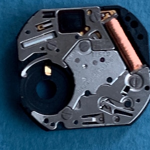 May include: Close-up of a watch movement with intricate metal components. The mechanism features silver-colored gears, a black circular element, and a copper-colored coil. The watch movement is set against a blue background.