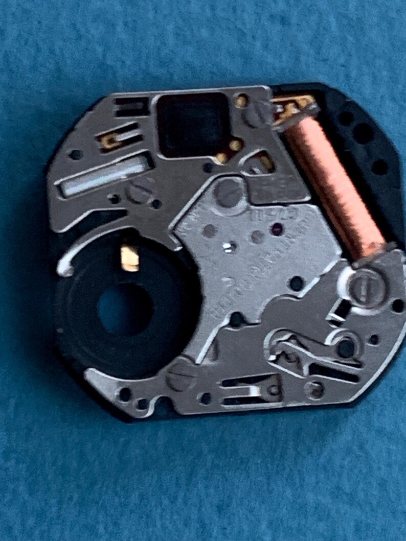 May include: Close-up of a watch movement with intricate metal components. The mechanism features silver-colored gears, a black circular element, and a copper-colored coil. The watch movement is set against a blue background.