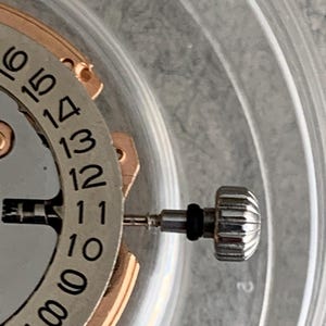 Puede incluir: Primer plano de un mecanismo de reloj con esfera plateada e indicador de fecha. La corona del reloj es plateada con dise&ntilde;o acanalado. Los n&uacute;meros del 7 al 16 son visibles en la esfera.