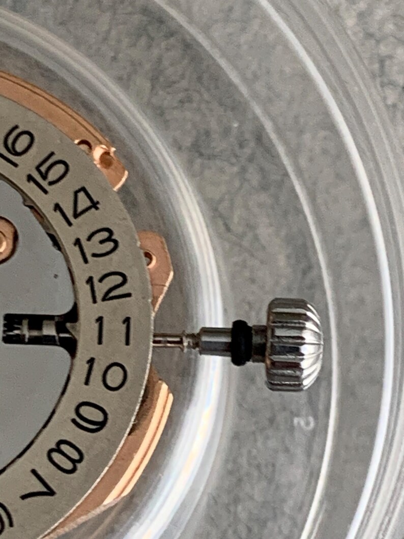 Puede incluir: Primer plano de un mecanismo de reloj con esfera plateada e indicador de fecha. La corona del reloj es plateada con dise&ntilde;o acanalado. Los n&uacute;meros del 7 al 16 son visibles en la esfera.