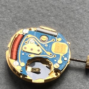 May include: Close-up of a watch movement with a gold-colored frame and a blue circuit board. The movement features a red coil, small screws, and the text "ZENITH" and "SWISS". The watch movement has seven jewels.