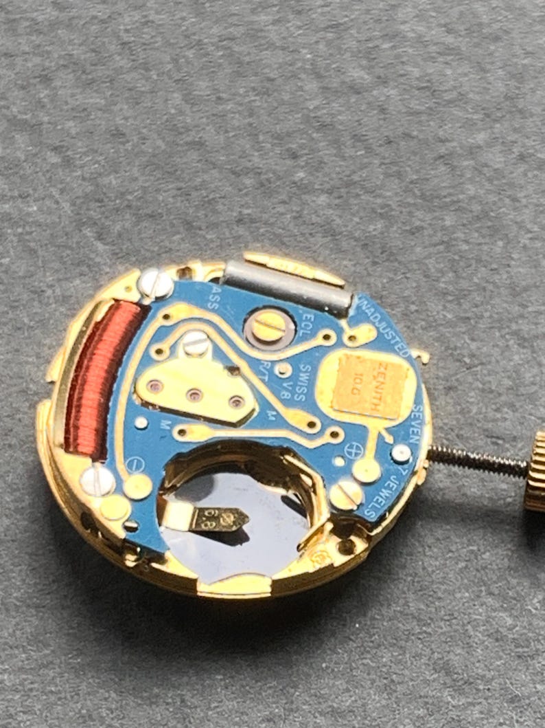 May include: Close-up of a watch movement with a gold-colored frame and a blue circuit board. The movement features a red coil, small screws, and the text "ZENITH" and "SWISS". The watch movement has seven jewels.