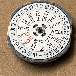 May include: A watch calendar dial with a white face, featuring black and red text indicating days of the week and dates. The dial is surrounded by a gray ring and has a central mechanism.