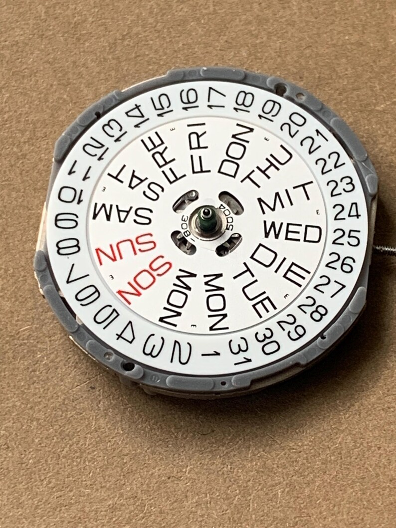 May include: A watch calendar dial with a white face, featuring black and red text indicating days of the week and dates. The dial is surrounded by a gray ring and has a central mechanism.
