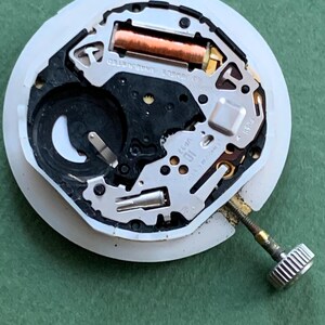 K&ouml;nnte beinhalten: Nahaufnahme eines Uhrwerks mit wei&szlig;er Kunststoffbasis. Es enth&auml;lt eine Kupferspule, eine Batterie und verschiedene Metallkomponenten. Eine silberne Krone ist angebracht. Der Text "NO JEWELS UNADJUSTED" ist sichtbar.
