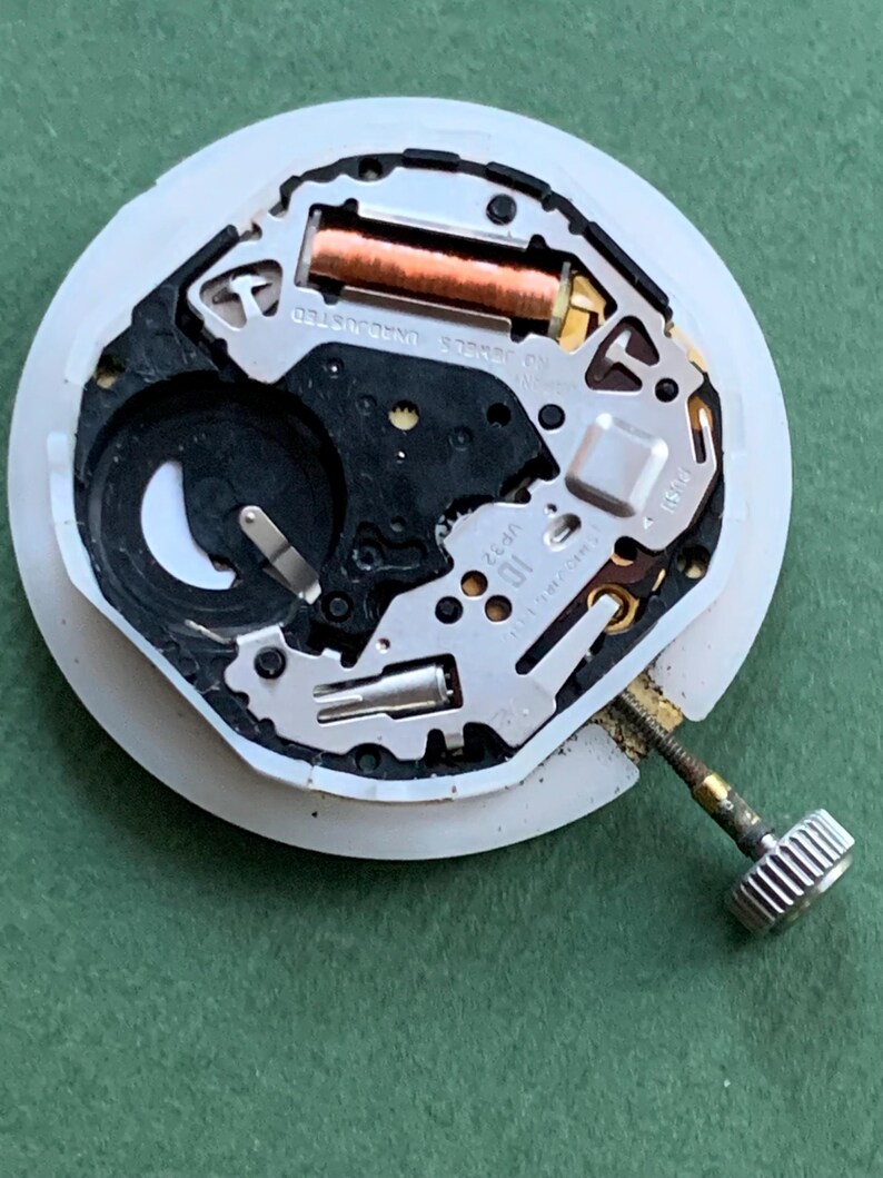 K&ouml;nnte beinhalten: Nahaufnahme eines Uhrwerks mit wei&szlig;er Kunststoffbasis. Es enth&auml;lt eine Kupferspule, eine Batterie und verschiedene Metallkomponenten. Eine silberne Krone ist angebracht. Der Text "NO JEWELS UNADJUSTED" ist sichtbar.