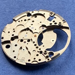Puede incluir: Primer plano de un movimiento de reloj plateado con numerosos agujeros y detalles intrincados. El componente circular tiene una secci&oacute;n curva y peque&ntilde;as joyas de colores. El fondo es azul liso.