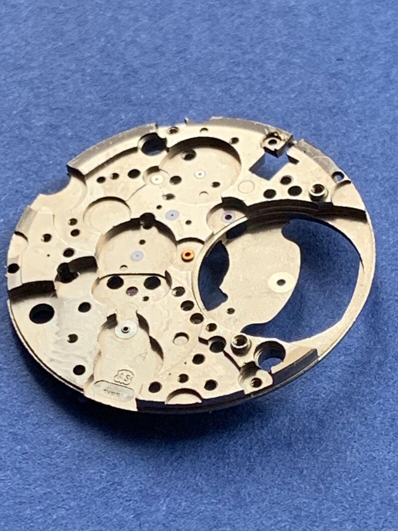 Puede incluir: Primer plano de un movimiento de reloj plateado con numerosos agujeros y detalles intrincados. El componente circular tiene una secci&oacute;n curva y peque&ntilde;as joyas de colores. El fondo es azul liso.