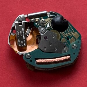 May include: A close-up of a watch movement on a red background. The movement features a green circuit board, metal components, and a black rubber piece. Text on a metal piece reads "ORE 1 JEWEL R790 H210 SWISS UNADJUSTED".