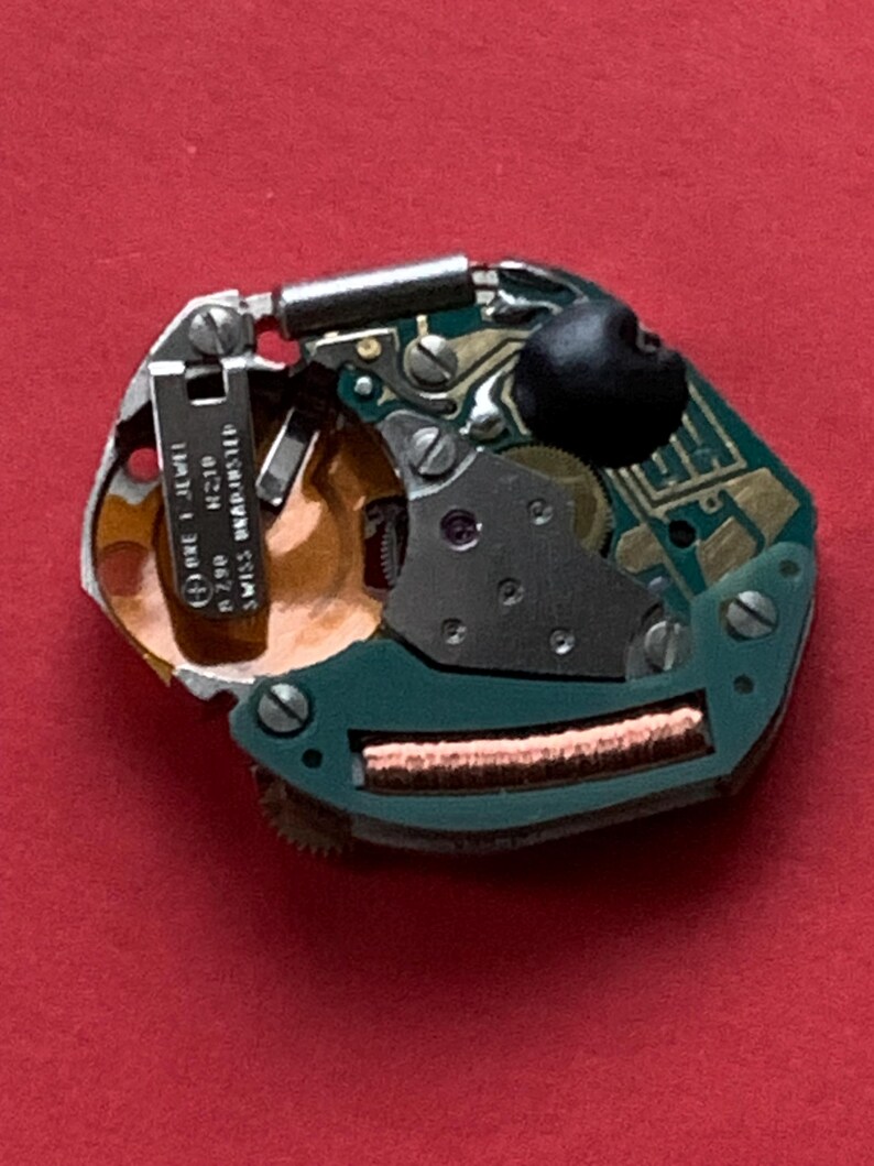 May include: A close-up of a watch movement on a red background. The movement features a green circuit board, metal components, and a black rubber piece. Text on a metal piece reads "ORE 1 JEWEL R790 H210 SWISS UNADJUSTED".