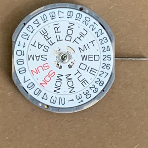 May include: A watch calendar dial with a white face and a gray metal frame. The days of the week are printed in black and red, with numbers 1-31 around the edge. A thin wire extends from the dial.