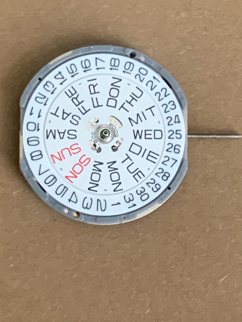 May include: A watch calendar dial with a white face and a gray metal frame. The days of the week are printed in black and red, with numbers 1-31 around the edge. A thin wire extends from the dial.