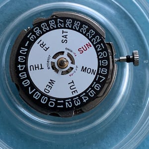 May include: A watch movement with a white dial showing days of the week and numbered dates. The dial has black numerals and text, with "SUN" in red. A small metal crown and other components are visible.