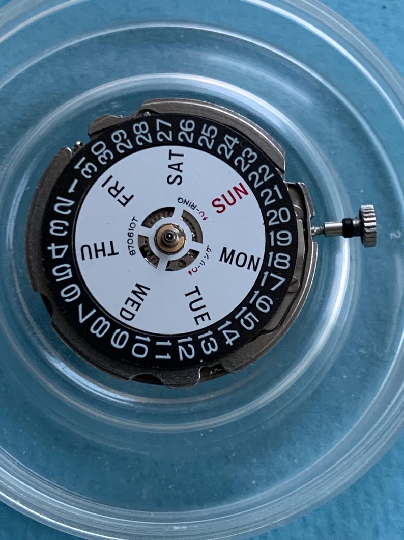 May include: A watch movement with a white dial showing days of the week and numbered dates. The dial has black numerals and text, with "SUN" in red. A small metal crown and other components are visible.