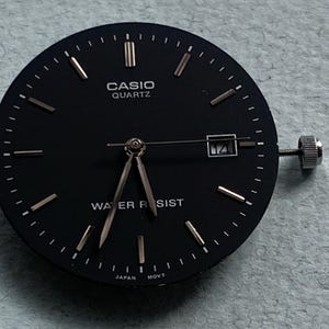 Puede incluir: Esfera de reloj Casio Quartz negra con agujas e indicadores dorados. La esfera muestra "CASIO QUARTZ" y "WATER RESIST". Ventana de fecha a las 3. Corona plateada.