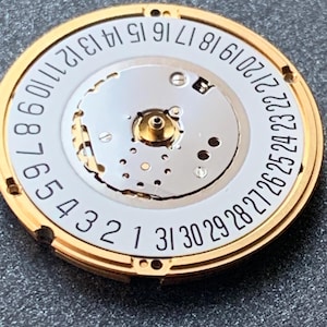 May include: Close-up of a watch movement with a gold-colored outer ring and a white inner dial. Black numbers from 1 to 31 are printed around the dial's edge. The watch mechanism is visible in the center.