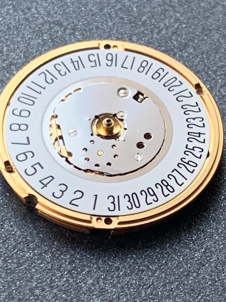 May include: Close-up of a watch movement with a gold-colored outer ring and a white inner dial. Black numbers from 1 to 31 are printed around the dial's edge. The watch mechanism is visible in the center.