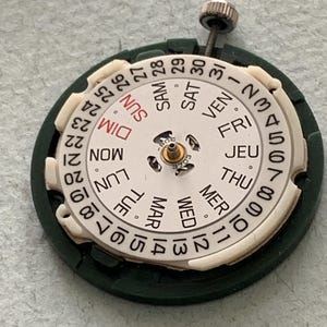 May include: A watch movement with a white dial and a dark green outer ring. The dial displays the days of the week in French and the numbers 1-31. A small screw is visible at the top. The watch part is designed for repair or restoration.