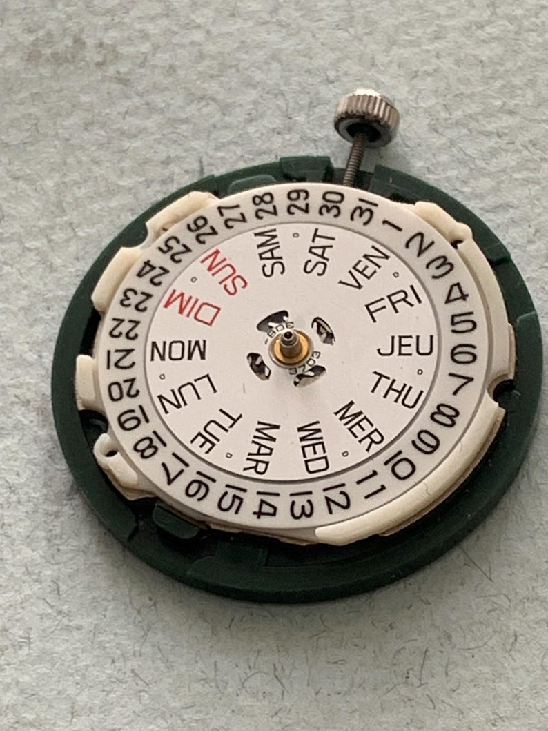 May include: A watch movement with a white dial and a dark green outer ring. The dial displays the days of the week in French and the numbers 1-31. A small screw is visible at the top. The watch part is designed for repair or restoration.