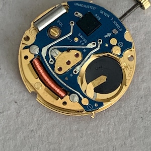 May include: A close-up of a watch movement with a gold-colored base and a blue circuit board. The board has the text "UNADJUSTED", "SEVEN 7 JEWELS", and "SWISS V8". A small battery and a copper coil are visible.
