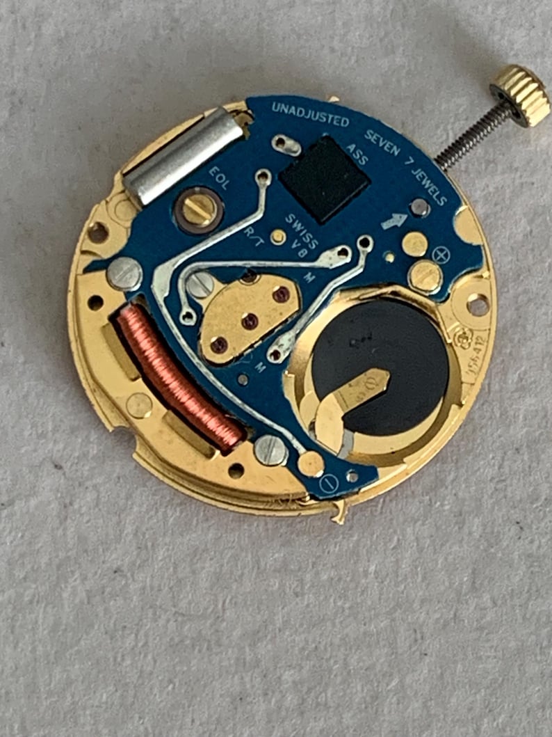 May include: A close-up of a watch movement with a gold-colored base and a blue circuit board. The board has the text "UNADJUSTED", "SEVEN 7 JEWELS", and "SWISS V8". A small battery and a copper coil are visible.