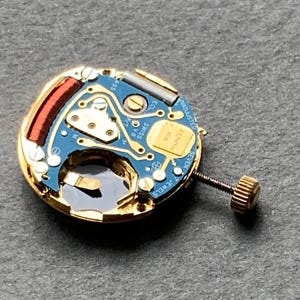 May include: Close-up of a watch movement with a blue circuit board and gold-colored components. The movement has a screw and a small gold-colored crown. The text "ZENITH" is visible on the movement.