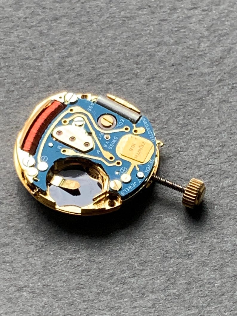 May include: Close-up of a watch movement with a blue circuit board and gold-colored components. The movement has a screw and a small gold-colored crown. The text "ZENITH" is visible on the movement.