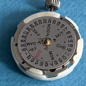 May include: A watch movement with a silver dial and a white plastic ring. The dial displays the days of the week and the numbers 1 through 31. A silver crown is at the top. The text includes "SON", "MON", "DIE", "FRE", and "SAM".