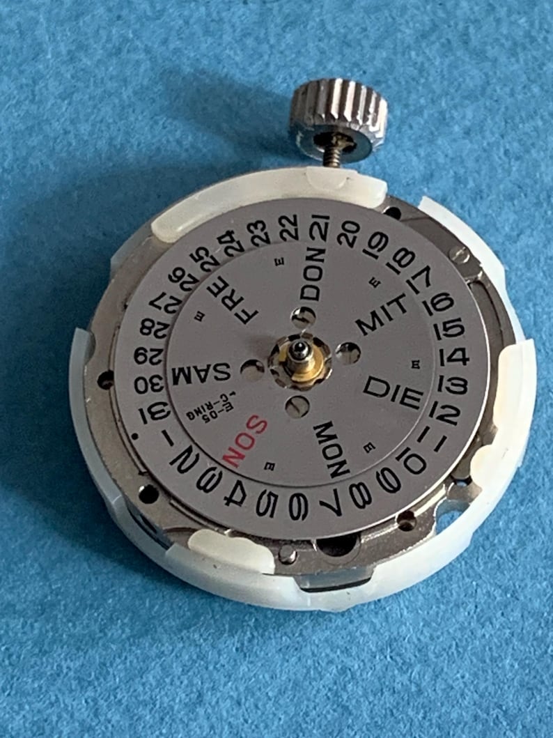 May include: A watch movement with a silver dial and a white plastic ring. The dial displays the days of the week and the numbers 1 through 31. A silver crown is at the top. The text includes "SON", "MON", "DIE", "FRE", and "SAM".