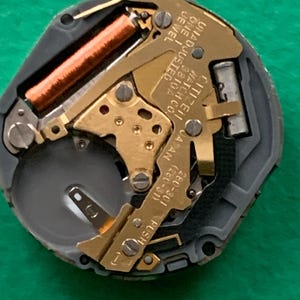 May include: Close-up of a watch movement with gold-colored gears, a copper-colored coil, and the text "CITIZEN WATCH CO. JAPAN". The watch mechanism is set against a gray and black frame.