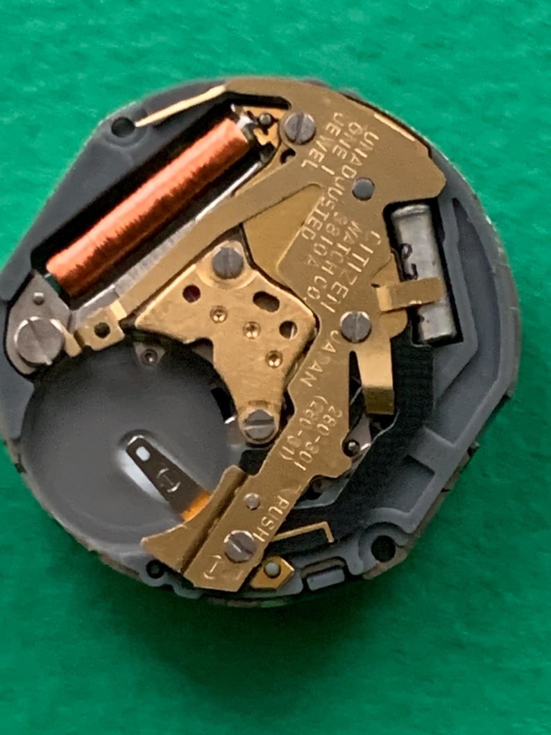 May include: Close-up of a watch movement with gold-colored gears, a copper-colored coil, and the text "CITIZEN WATCH CO. JAPAN". The watch mechanism is set against a gray and black frame.