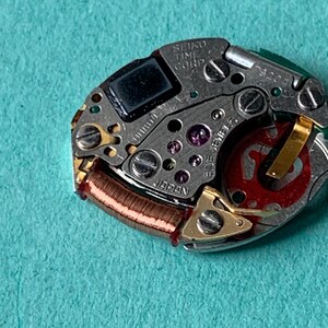 May include: Close-up of a Seiko watch movement. The intricate mechanism features silver metal components, including screws, gears, and a red rotor. The text "SEIKO TIME CORP" and "JAPAN" are visible. The background is a teal color.