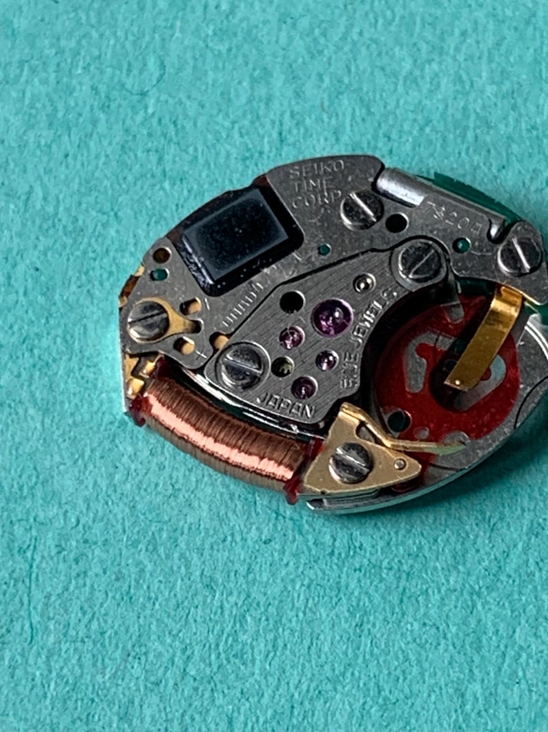 May include: Close-up of a Seiko watch movement. The intricate mechanism features silver metal components, including screws, gears, and a red rotor. The text "SEIKO TIME CORP" and "JAPAN" are visible. The background is a teal color.