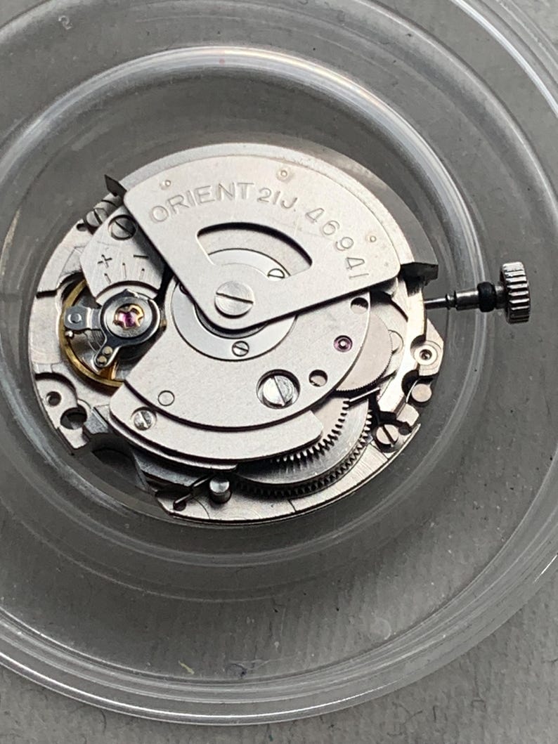 Pu&ograve; includere: Primo piano di un movimento di orologio argentato con la scritta "ORIENT 21J 46941". L'intricato meccanismo include piccoli ingranaggi, viti e gioielli. Una corona &egrave; visibile a destra. Il movimento dell'orologio &egrave; all'interno di un contenitore trasparente.