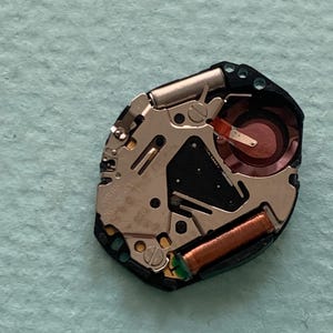 May include: A detailed view of a watch movement, revealing its complex internal mechanisms. The movement includes silver metal components, a copper coil, and a red circular element. The black plastic base houses the internal parts.