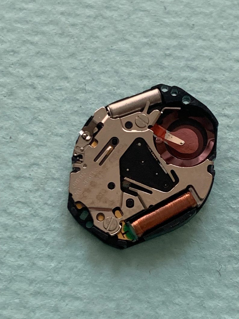 May include: A detailed view of a watch movement, revealing its complex internal mechanisms. The movement includes silver metal components, a copper coil, and a red circular element. The black plastic base houses the internal parts.