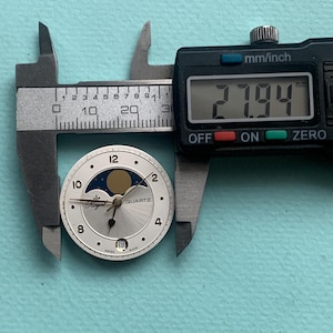 Op de afbeelding: Een zilveren Royal Quartz wijzerplaat met een maanfase-ontwerp, gemeten op 27,94 mm met een digitale schuifmaat. De wijzerplaat heeft zwarte cijfers en wijzers, met een datumweergave op 6 uur. De achtergrond is lichtblauw.
