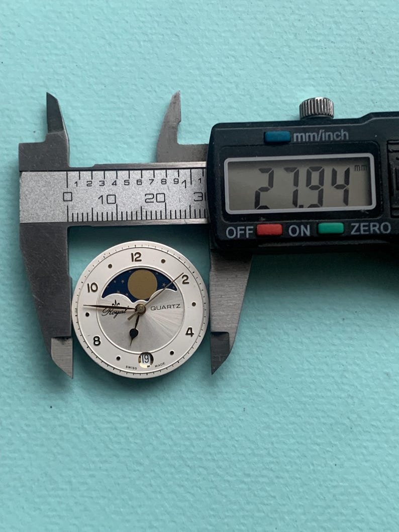 Op de afbeelding: Een zilveren Royal Quartz wijzerplaat met een maanfase-ontwerp, gemeten op 27,94 mm met een digitale schuifmaat. De wijzerplaat heeft zwarte cijfers en wijzers, met een datumweergave op 6 uur. De achtergrond is lichtblauw.