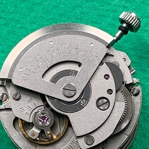May include: Close-up of a silver Orient watch movement with visible gears, jewels, and the text "ORIENT WATCH CO. LTD." and "TWENTY-ONE JEWELS JAPAN". A silver crown is visible at the top. The background is green.