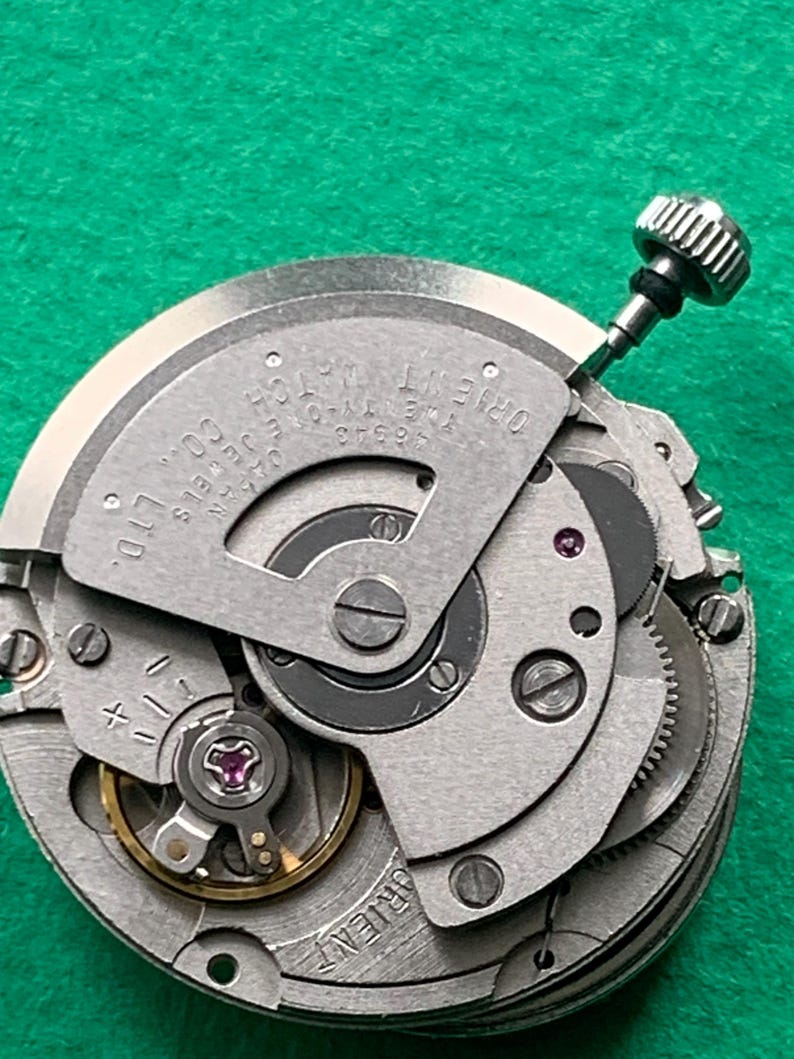 May include: Close-up of a silver Orient watch movement with visible gears, jewels, and the text "ORIENT WATCH CO. LTD." and "TWENTY-ONE JEWELS JAPAN". A silver crown is visible at the top. The background is green.